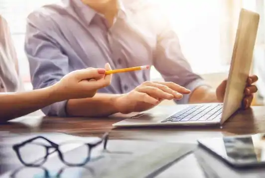 Woman pointing with pencil at a laptop, while working with her husband at a table with glasses and documents on it.