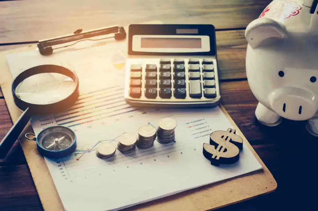 A magnifying glass, compass, quarters in stacks, calculator, and dollar bill sign on documents with graphs on a clipboard, next to a piggy bank on a table.
