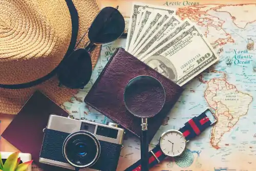 A hat, sunglasses, passport, camera, watch, magnifying glass, and wallet with multiple one-hundred dollar bills in it, on a large world map.