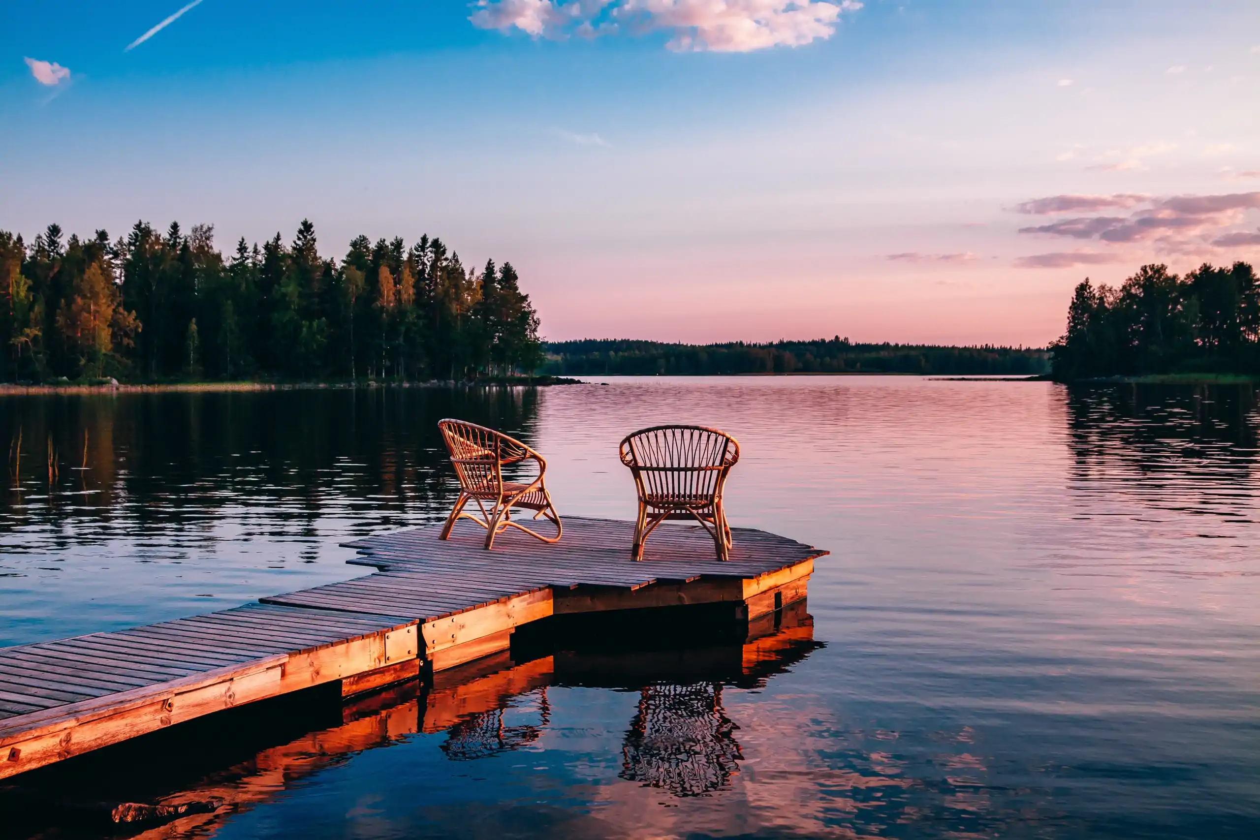 Two wooden chairs at the end of a dock in the middle of a lake during sunset.